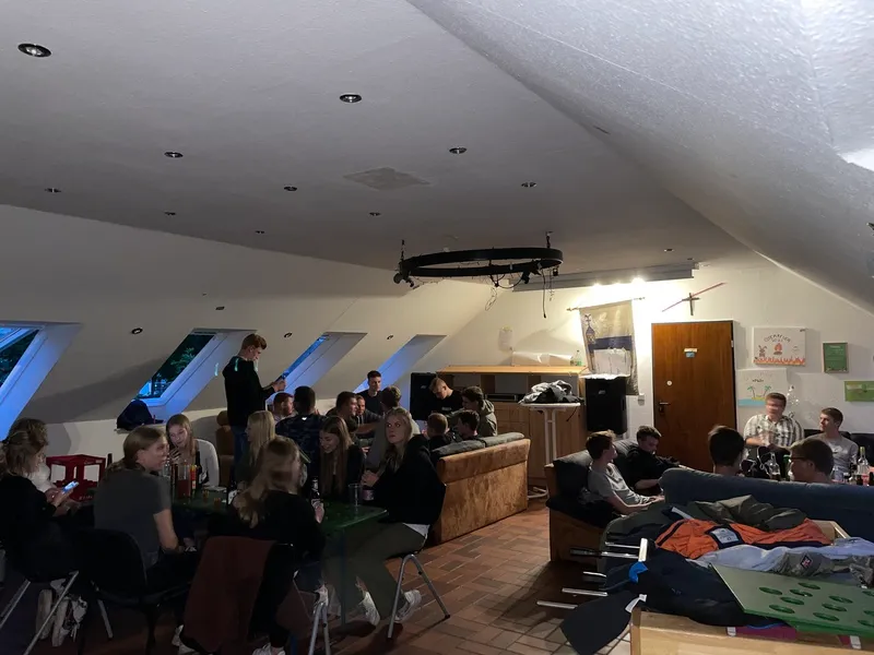 Eine größere Gruppe von Menschen sitzt und unterhält sich in einem gemütlichen Dachgeschossraum mit schräger Decke, Dachfenstern, Sofas und Tischen.