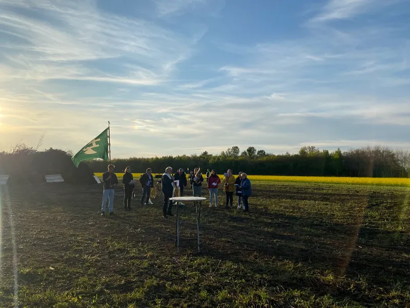 Eine Gruppe von Menschen steht bei tief stehender Sonne auf einem Acker neben einem kleinen Tisch, im Hintergrund weht eine grüne Fahne mit weißem Symbol vor einem gelben Feld und einer Baumreihe.